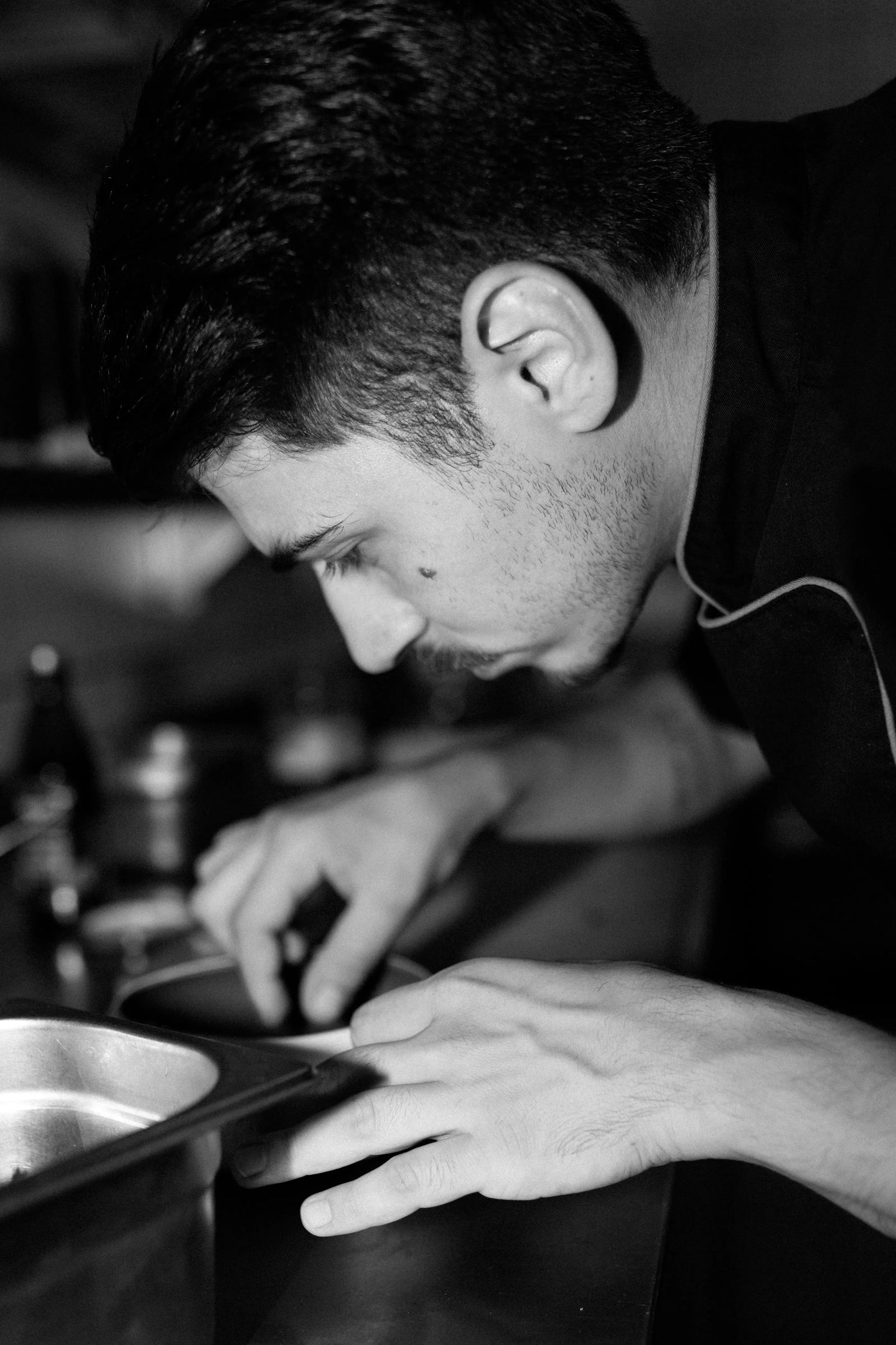 Schwarz-Weiß-Foto im Restaurant Buonissimo: Ein Nahaufnahme-Profilbild von Koch Giuseppe, der sich tief über eine Arbeitsfläche beugt. Er trägt eine dunkle Kochjacke und konzentriert sich fokussiert auf das Anrichten einer Speise mit beiden Händen. Im Vordergrund ist eine Metallschale unscharf zu erkennen.