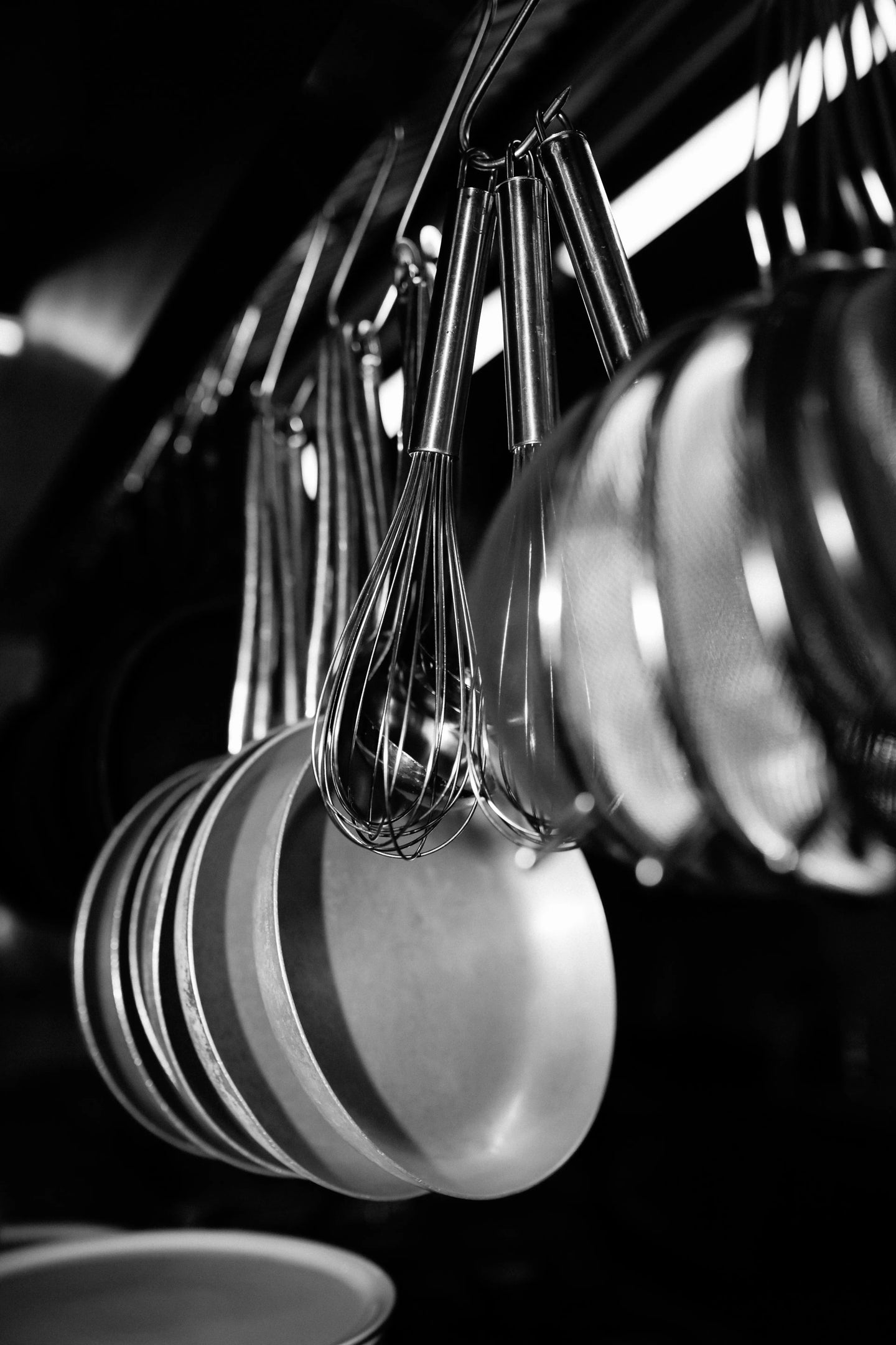 Schwarz-Weiß-Aufnahme von Küchenutensilien im Buonissimo Neu-Ulm: Mehre Schneebesen aus Edelstahl und eine Reihe kleiner Pfannen hängen griffbereit an Metallhaken in der Restaurantküche. Das Spiel aus Licht und Schatten hebt die metallischen Texturen hervor.