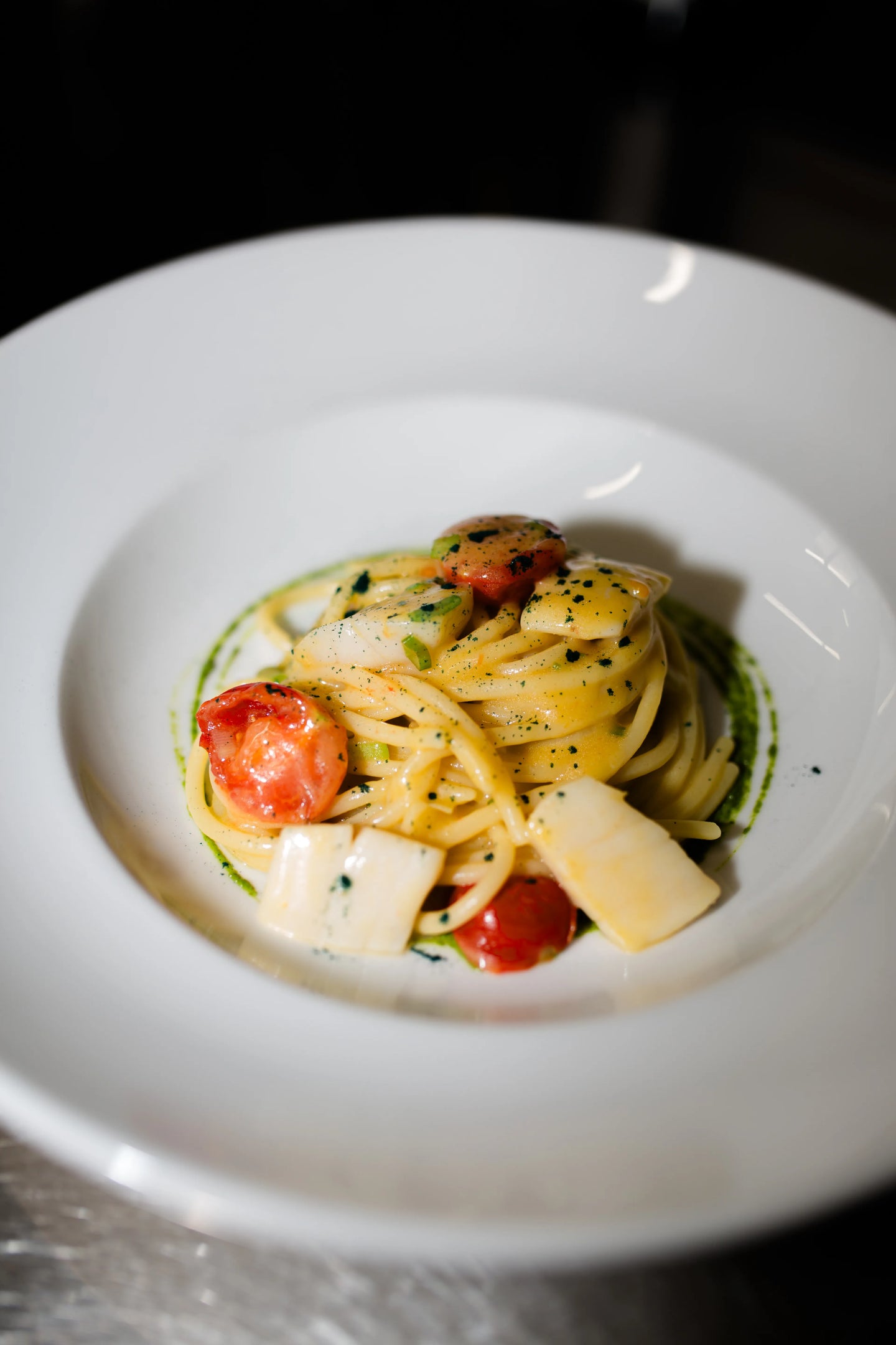 Feines Pasta-Gericht aus dem Restaurant Buonissimo Neu-Ulm: Ein elegant gedrehter Nest-Haufen Spaghetti, garniert mit hellem Tintenfisch, halbierten Kirschtomaten und feinen Kräutern. Das Gericht ist in einem tiefen, weißen Teller serviert und von einem Dekoring aus grünem Kräuteröl umrahmt.